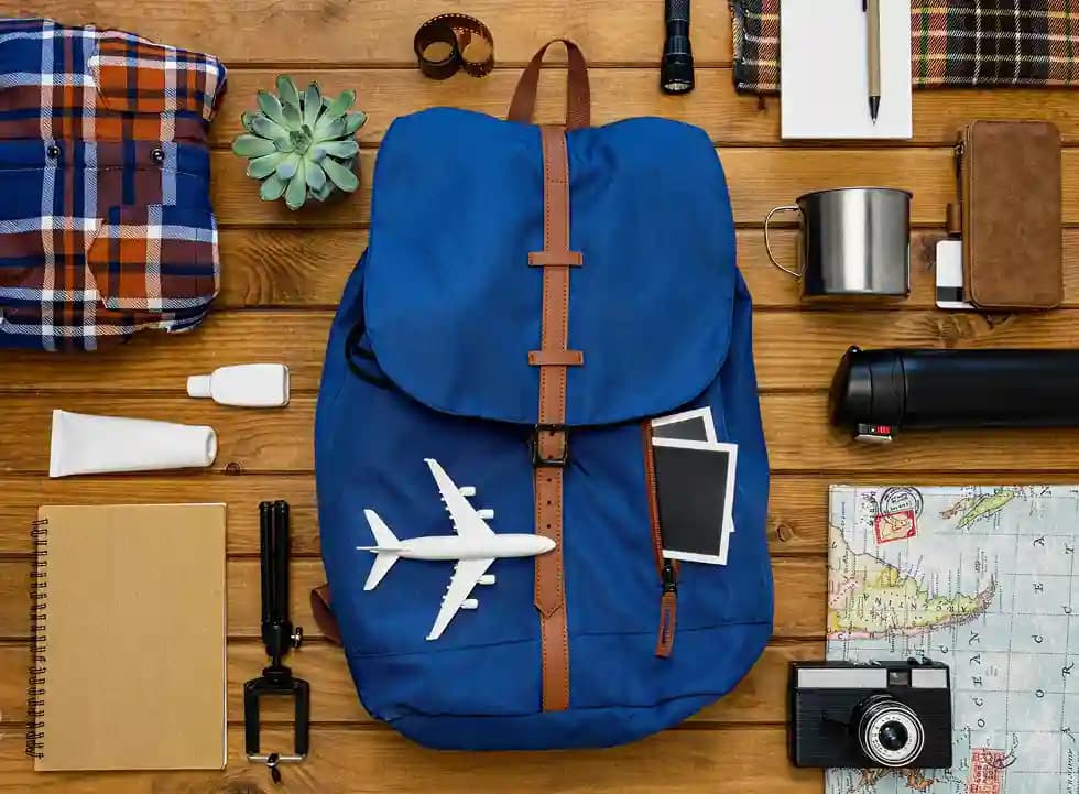 A backpack surrounded by travel essentials laid out on a wooden table