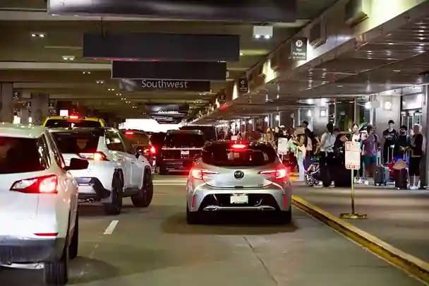 Cars queued along a busy airport departures curb at night as travelers wait on the sidewalk