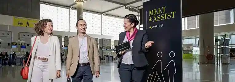An airport greeter walking two travelers through a terminal past a Meet and Assist stand